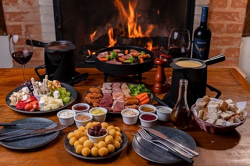 Mesa farta de fondue com carnes na chapa, queijos, frutas, pães e molhos, ao lado da lareira