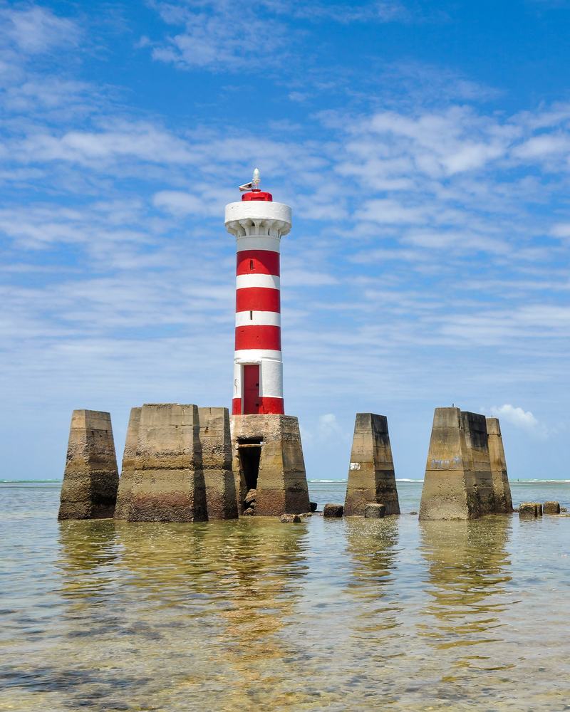 Farol da Ponta Verde, listrado de vermelho e branco, dentro do mar raso de Maceió na maré baixa